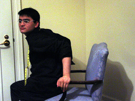 Josh tests the posing chair in the corner of the room.   Measuring tape is wrapped around his neck.